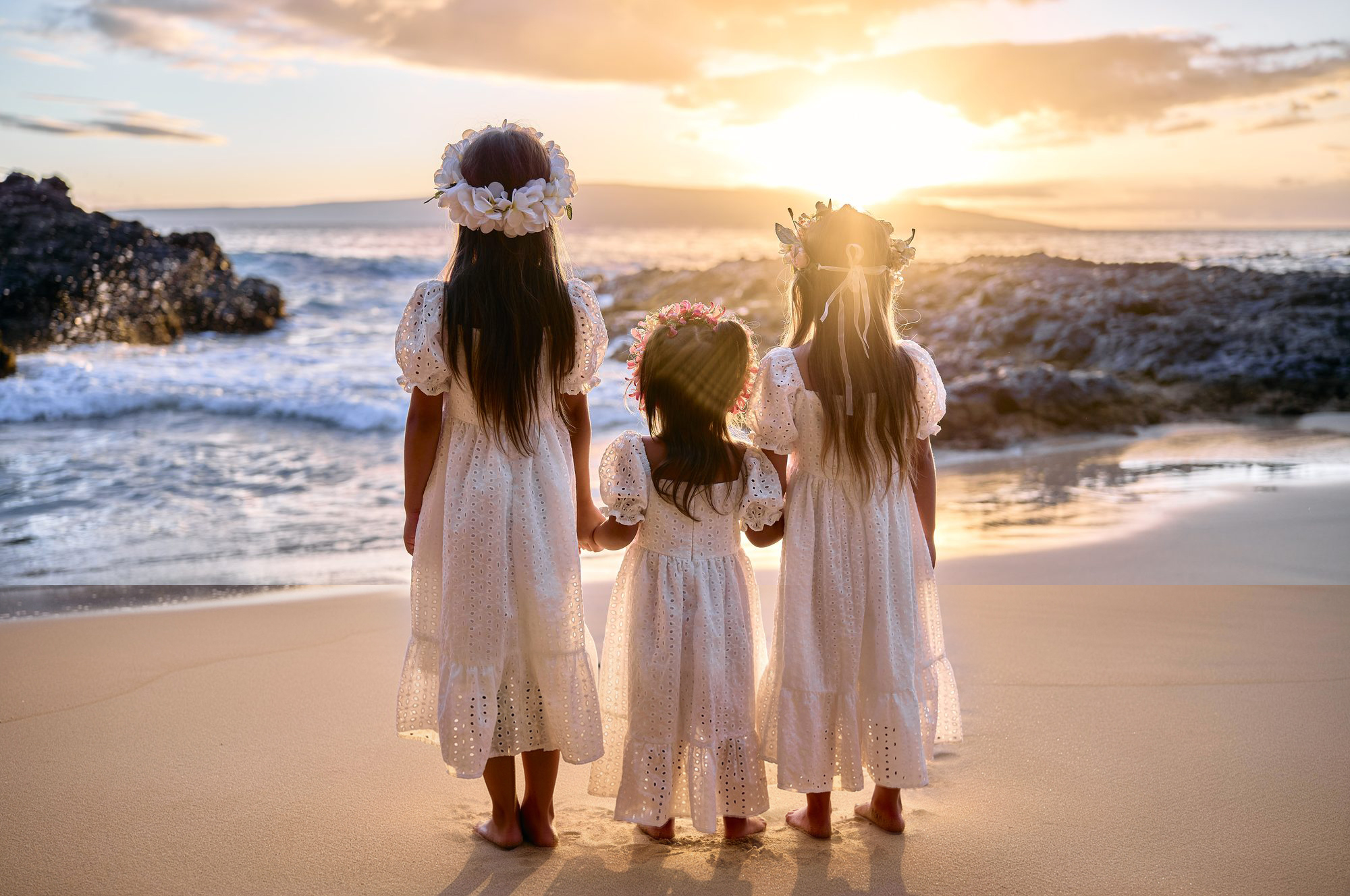 Three sisters in white flower crowns facing a Maui sunset, backlit by direct sun