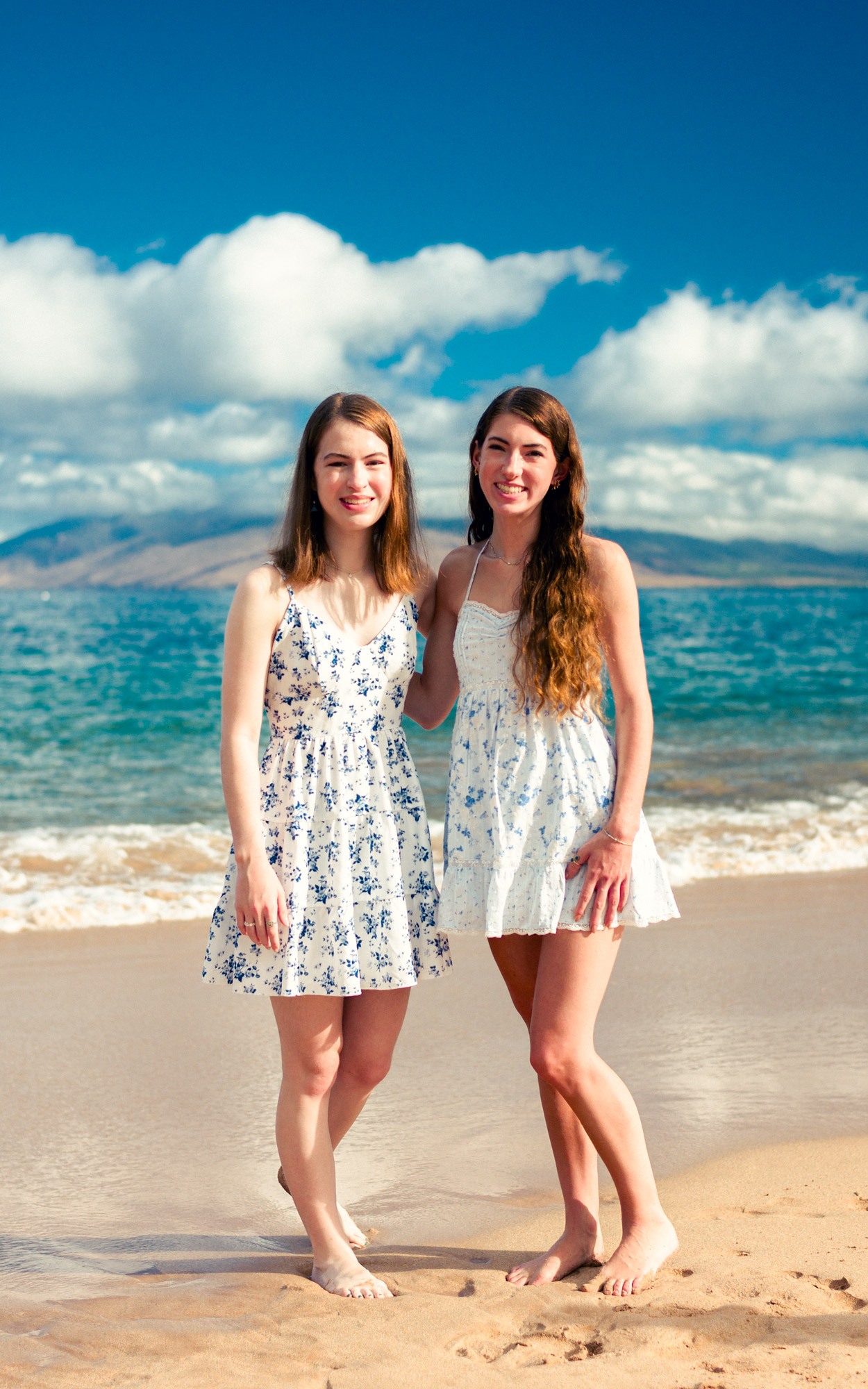 Two sisters in floral dresses on the sand with turquoise water behind them in Maui - image by Wailea Photo®