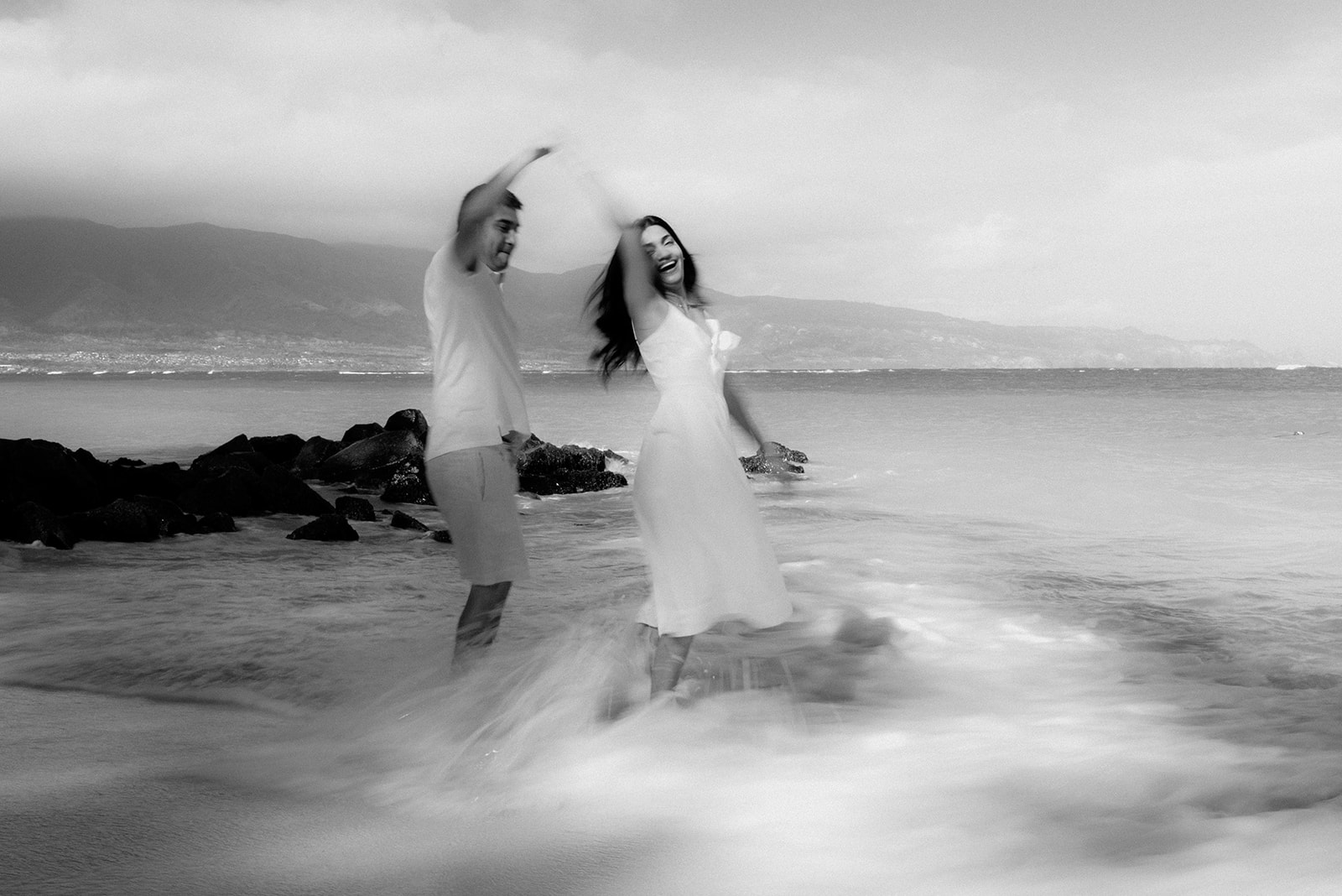 Laughing couple dancing in the surf beside lava rocks, motion-blurred in black and white