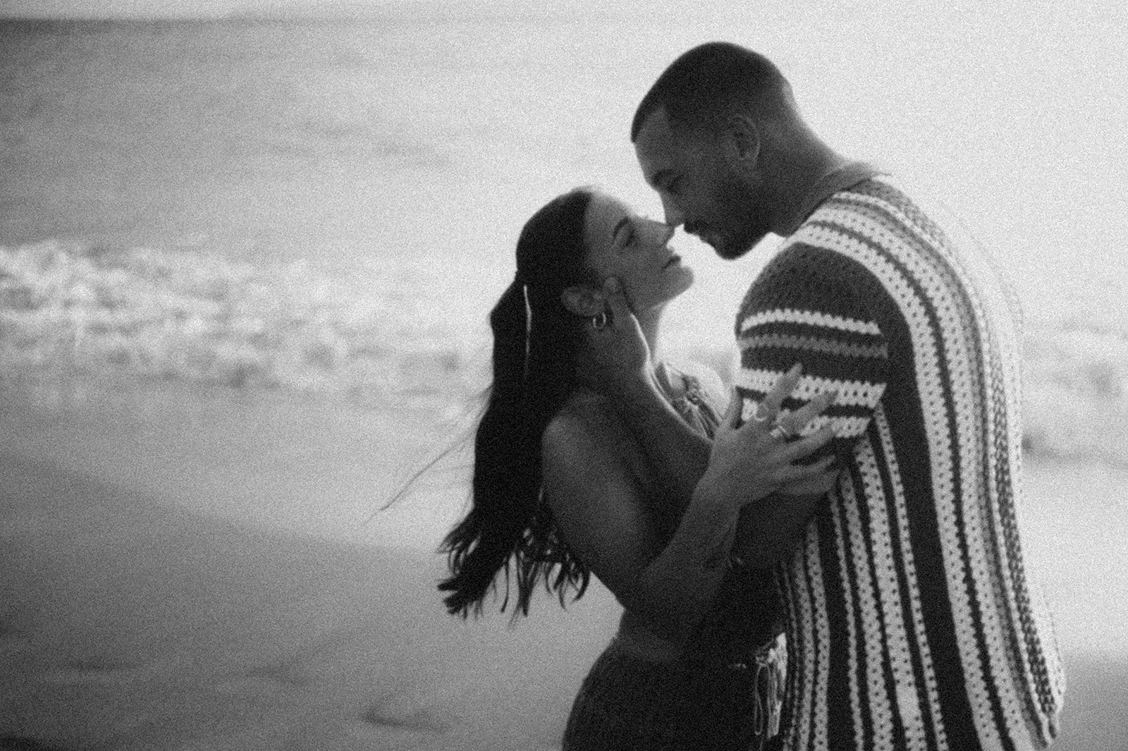 Couple nose to nose at the water’s edge, her hand on his cheek, in black and white