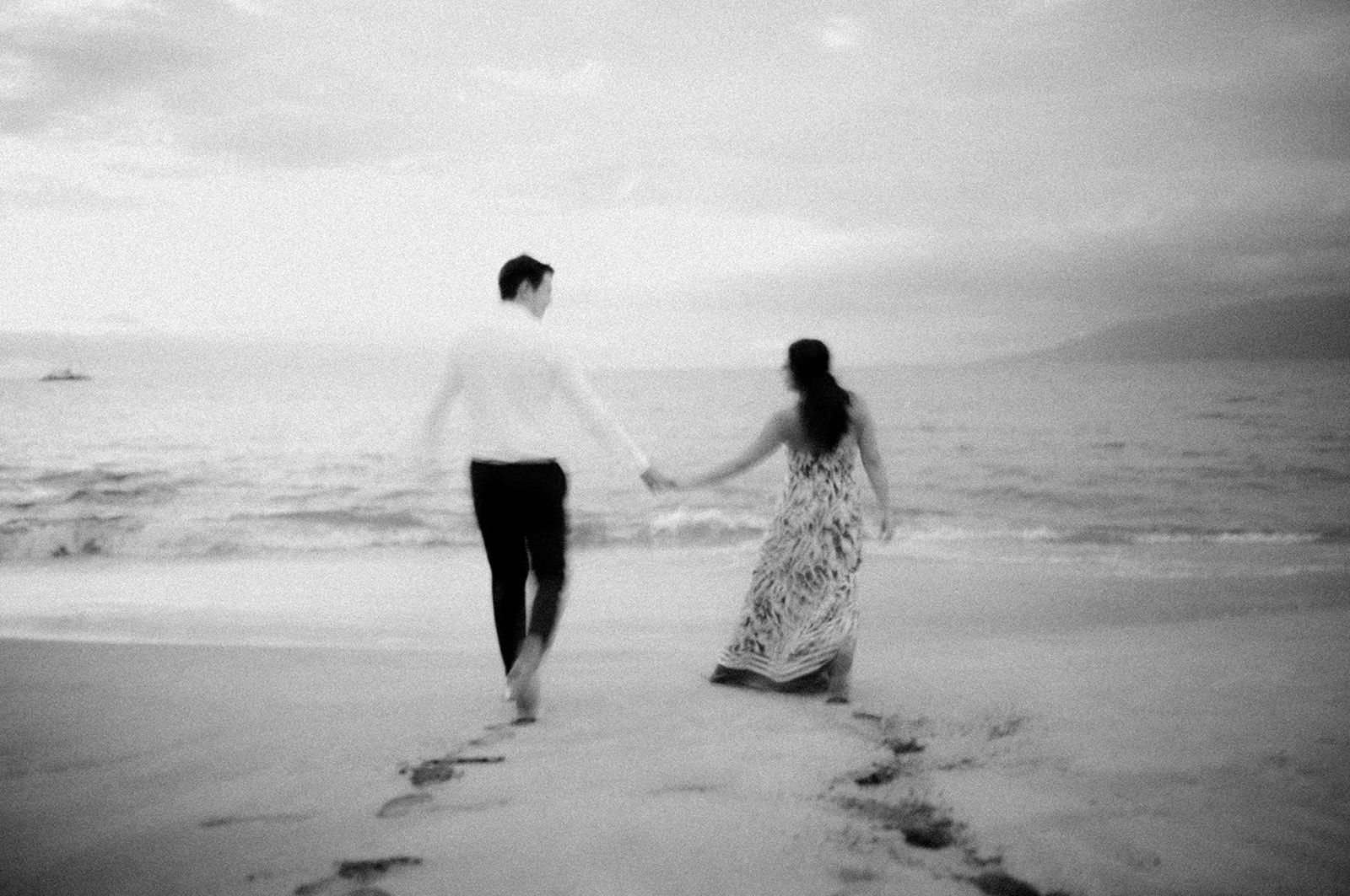 Couple walking hand in hand toward the sea, footprints trailing in the sand, in black and white