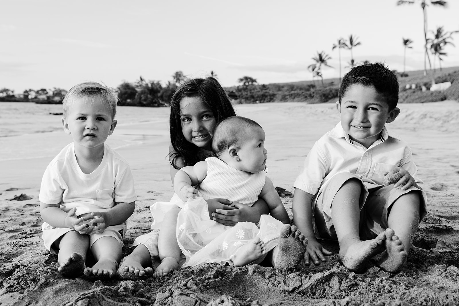 Four young children sitting barefoot in the sand with palms behind, in black and white