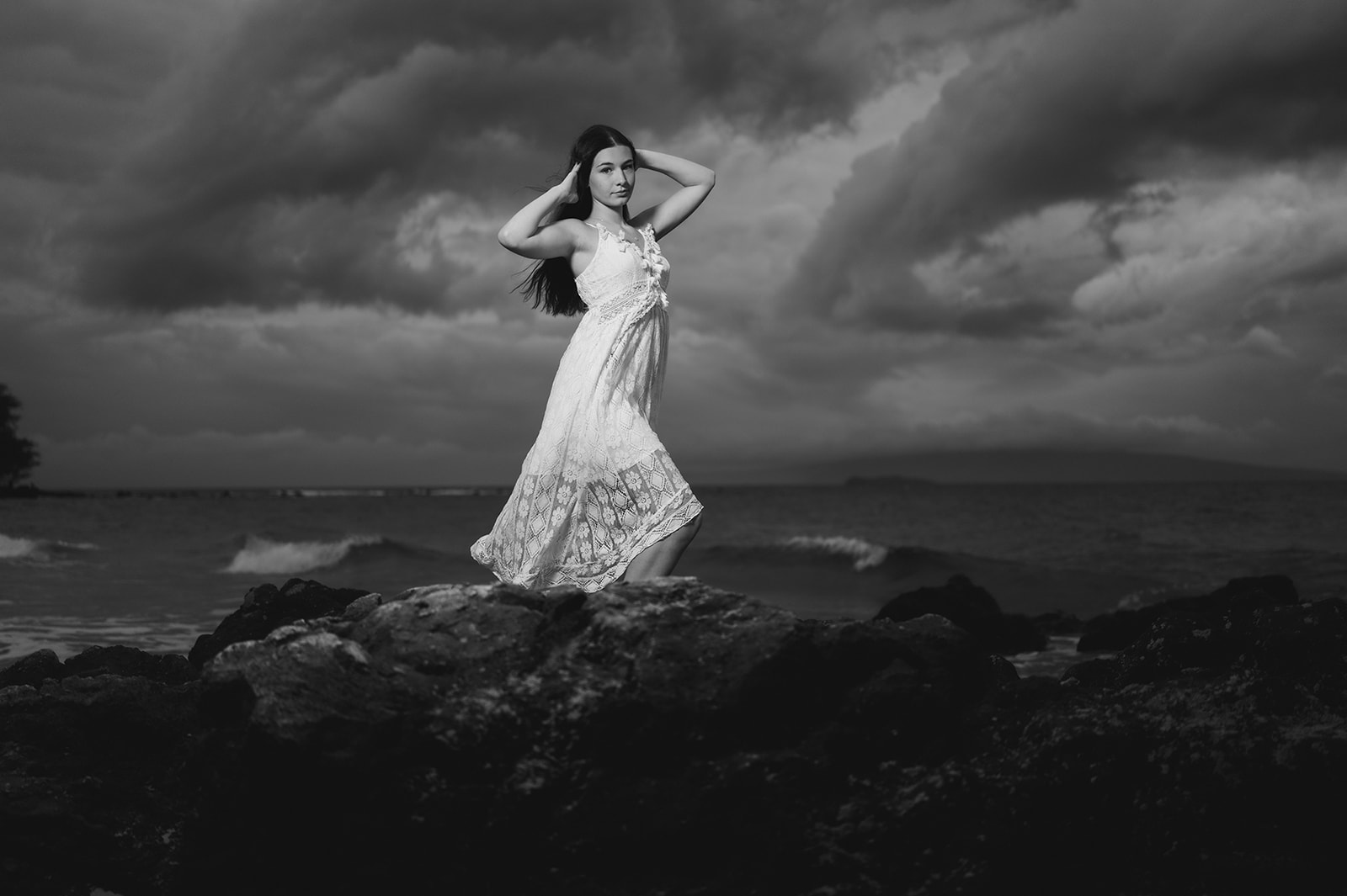Young woman in a white lace dress on lava rock beneath dramatic storm clouds, in black and white