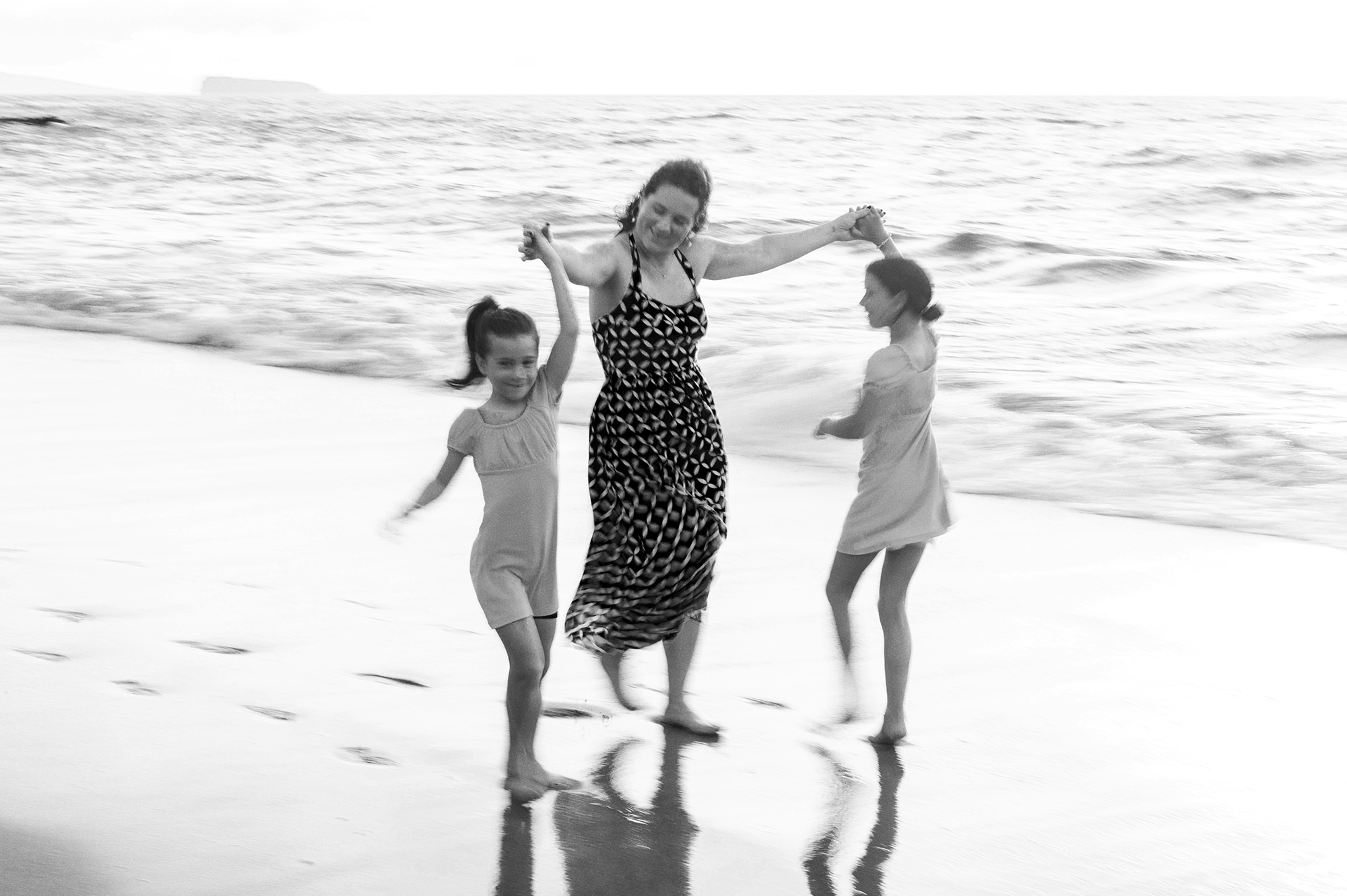 Mother dancing hand in hand with her two daughters at the water’s edge, Molokini on the horizon, in black and white