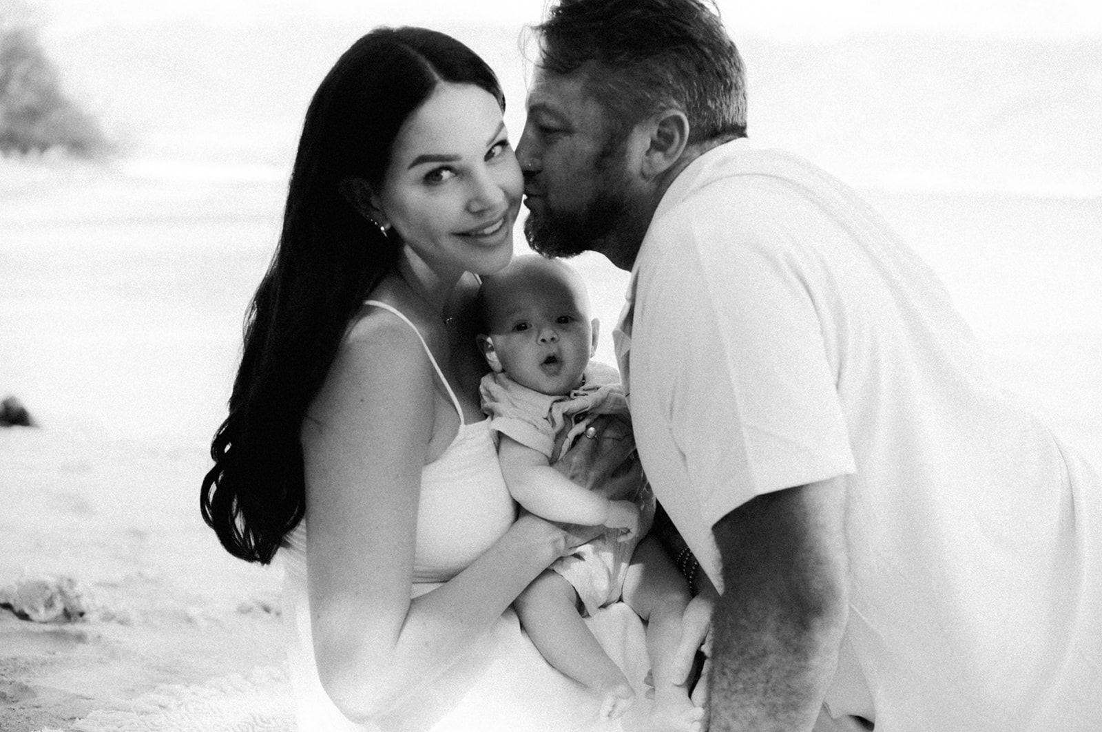 Father kissing mother’s cheek as she holds their wide-eyed baby on the beach, in black and white