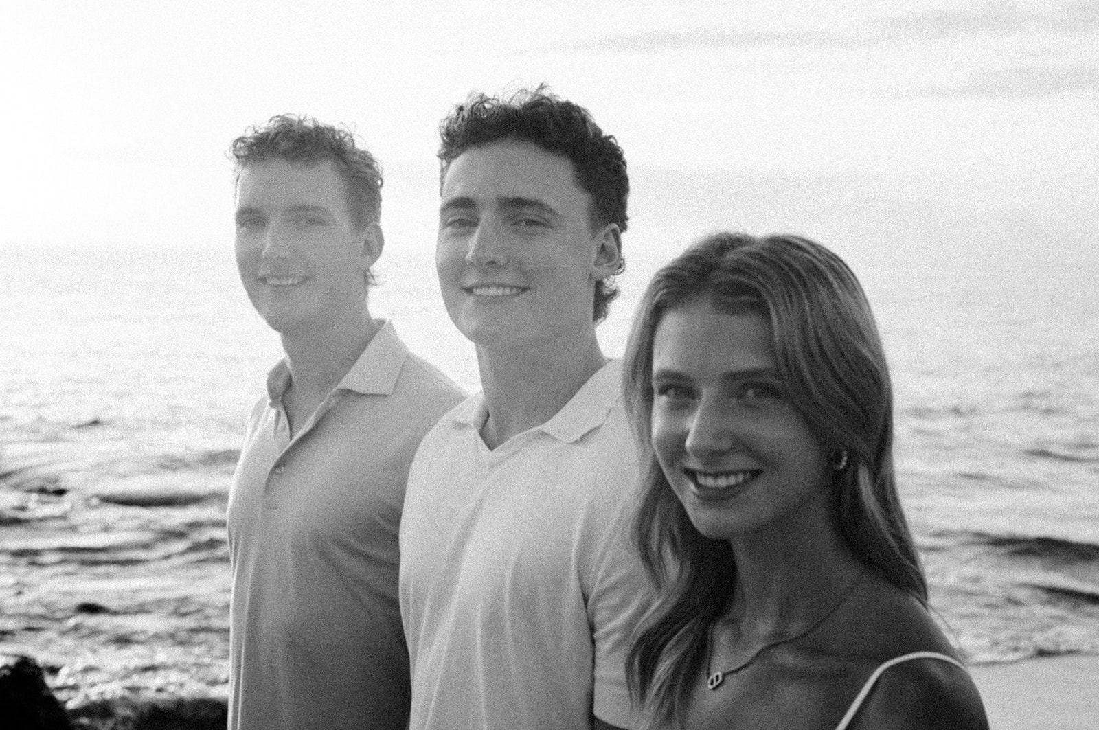 Three grown siblings smiling over their shoulders with the ocean behind, in black and white