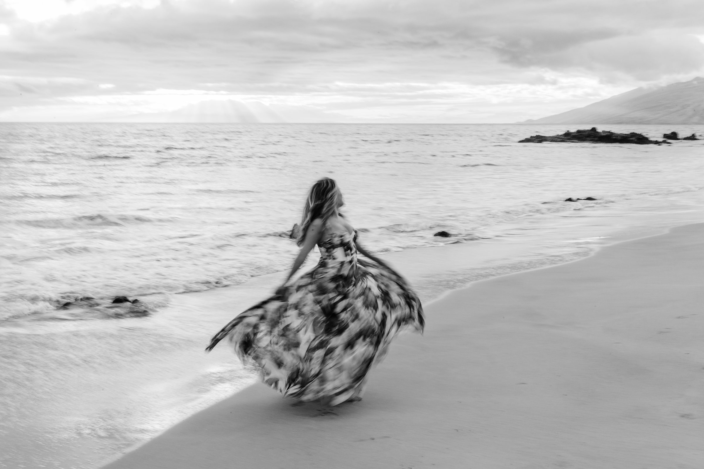 Woman twirling in a flowing patterned gown at the water’s edge, West Maui mountains beyond, in black and white
