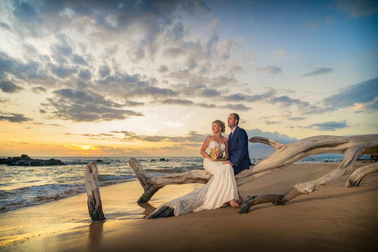 A newly married couple seated on driftwood beside the ocean at sunset in Maui