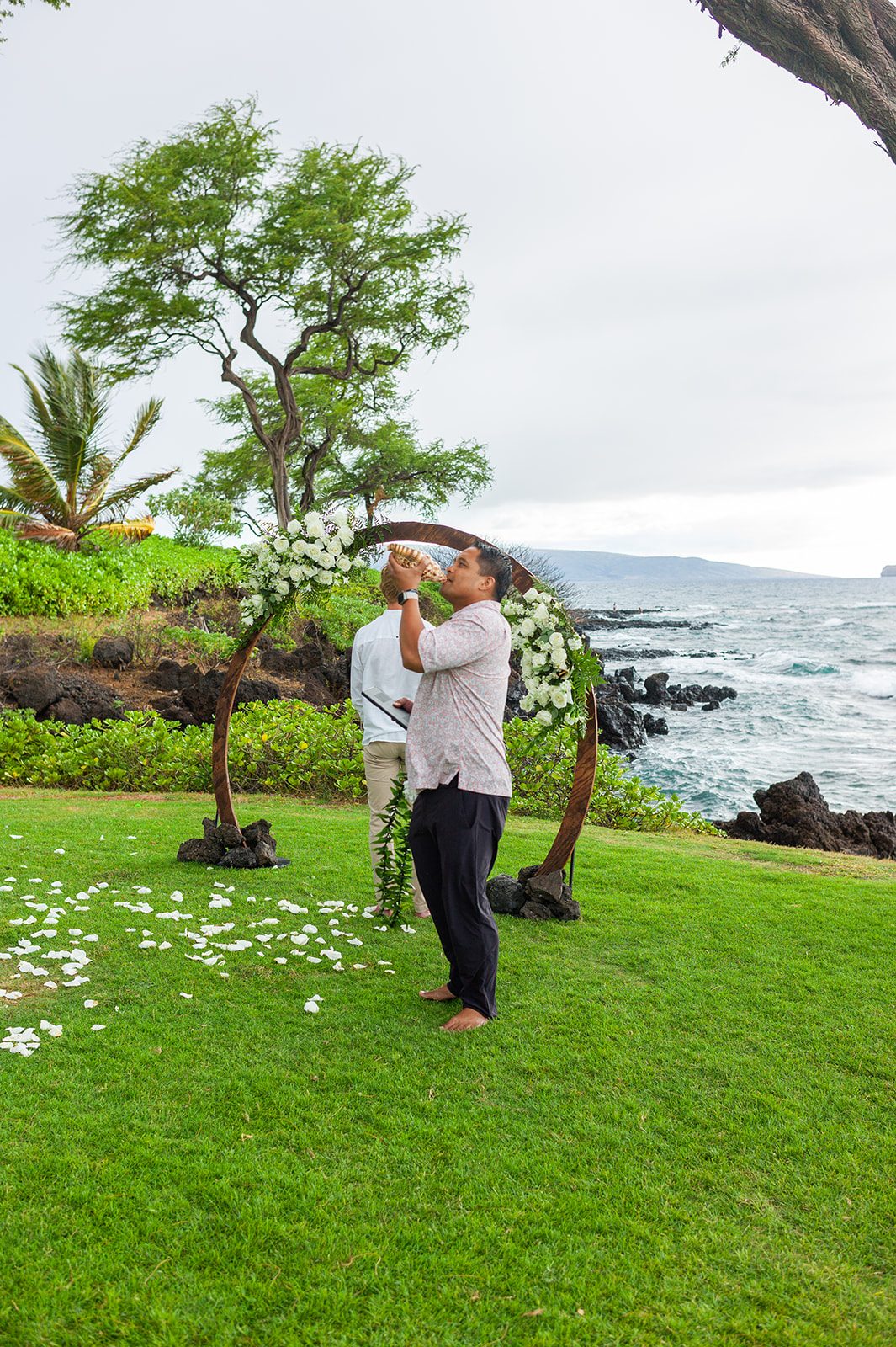 Wedding officiant Derick Sebastian blowing a Hawaiian conch shell, or pū, beside the ocean before a Maui wedding ceremony