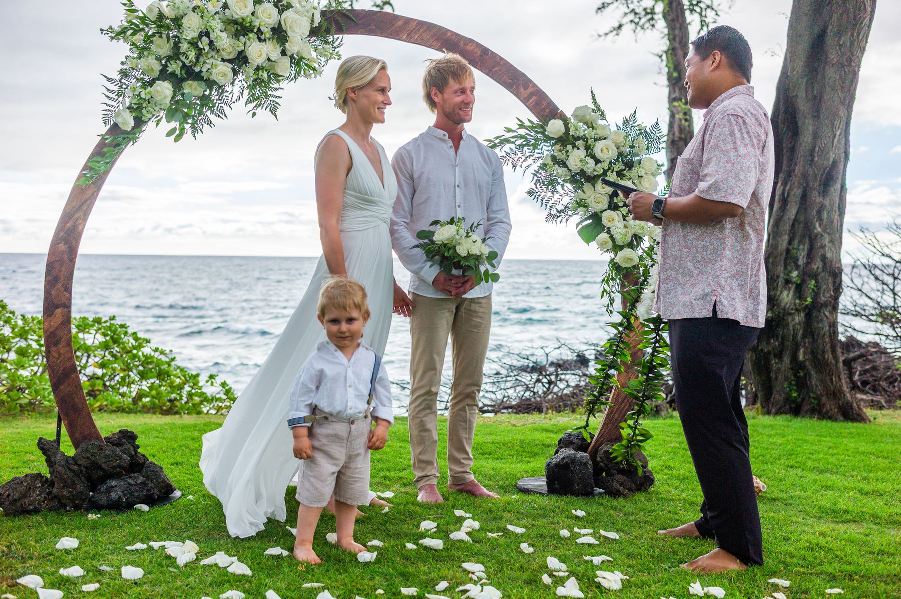 Wedding officiant Derick Sebastian leading an oceanfront ceremony in Maui as a couple and their young son stand beneath a floral arch