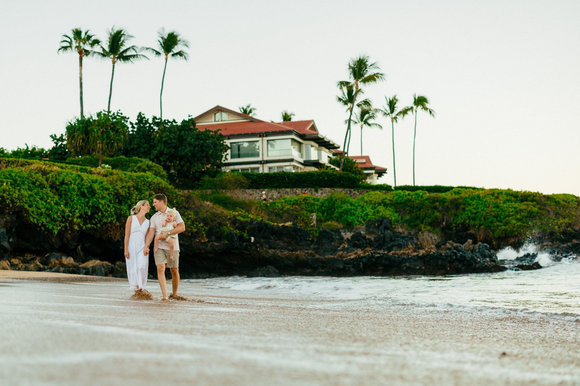 A family walking the shoreline at first light below the hillside homes and palms at Four Seasons Beach in Wailea