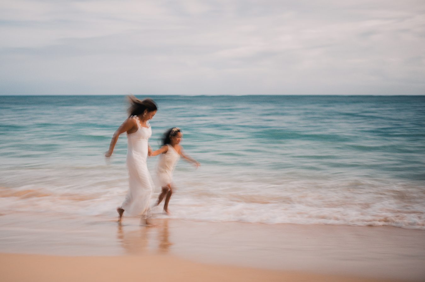 Una madre y su hija con vestidos blancos caminando de la mano por la orilla de Maui, con el movimiento difuminado