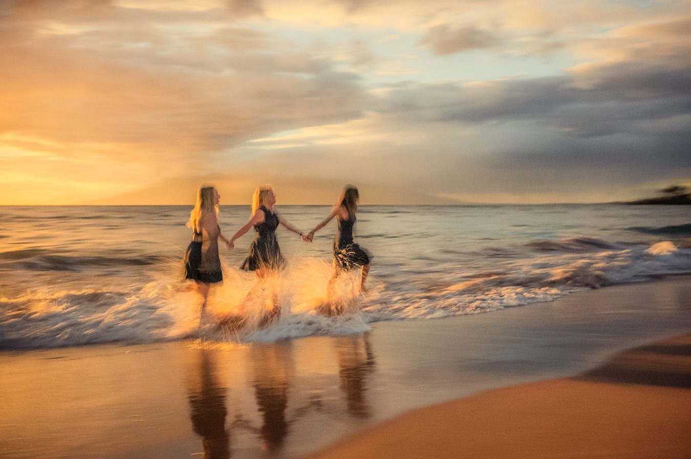 Tres amigas tomadas de la mano corriendo por el oleaje al atardecer en Maui, con un desenfoque pictórico