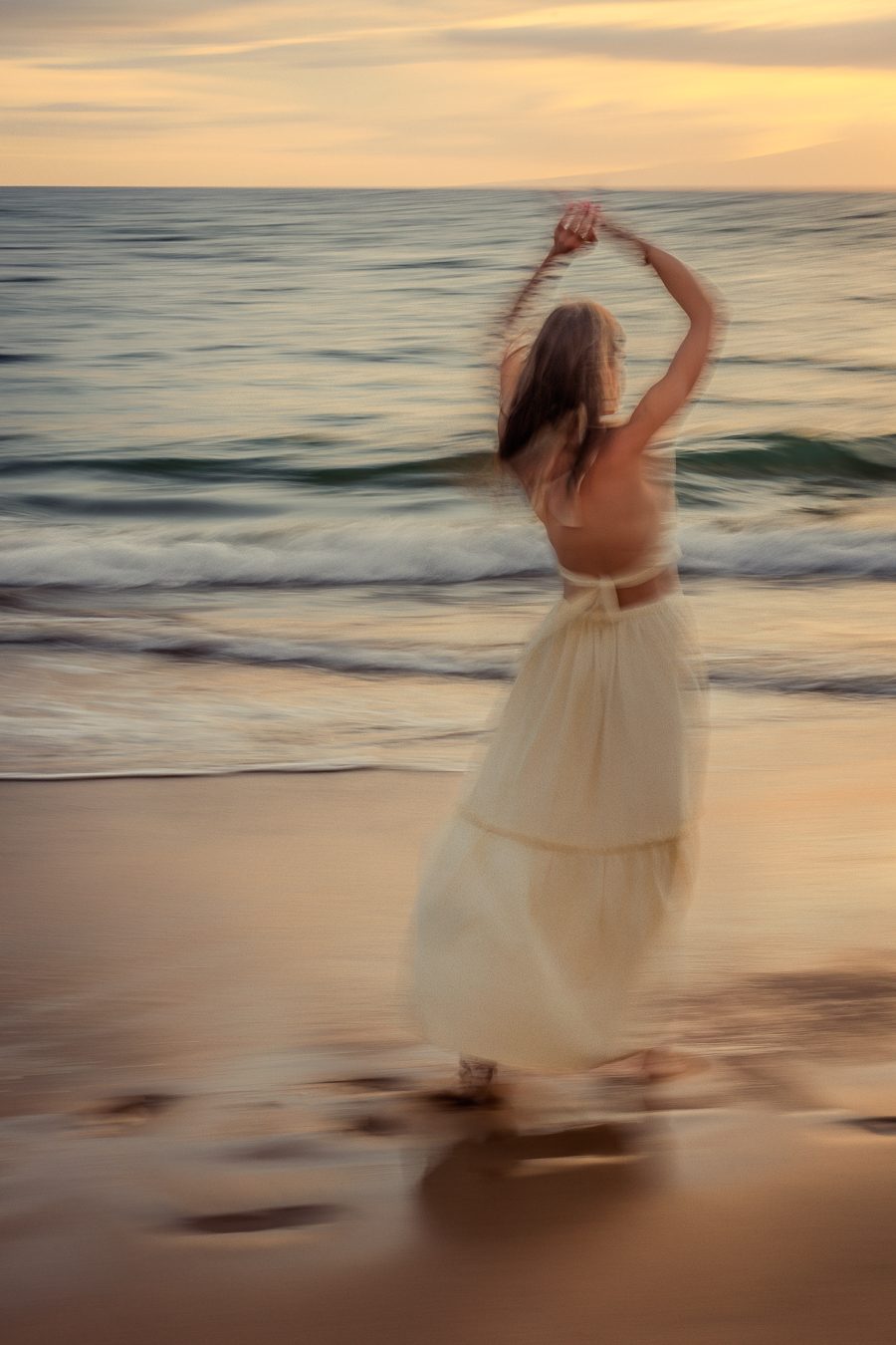 Mujer con vestido color crema girando con los brazos en alto en una playa de Maui al atardecer, desenfoque pictórico de movimiento
