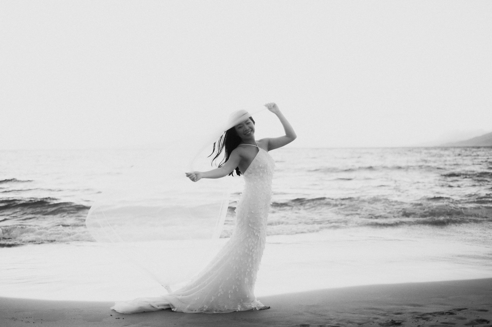 Black and white portrait of a bride in a sunhat with her veil blowing in the wind on a Maui beach