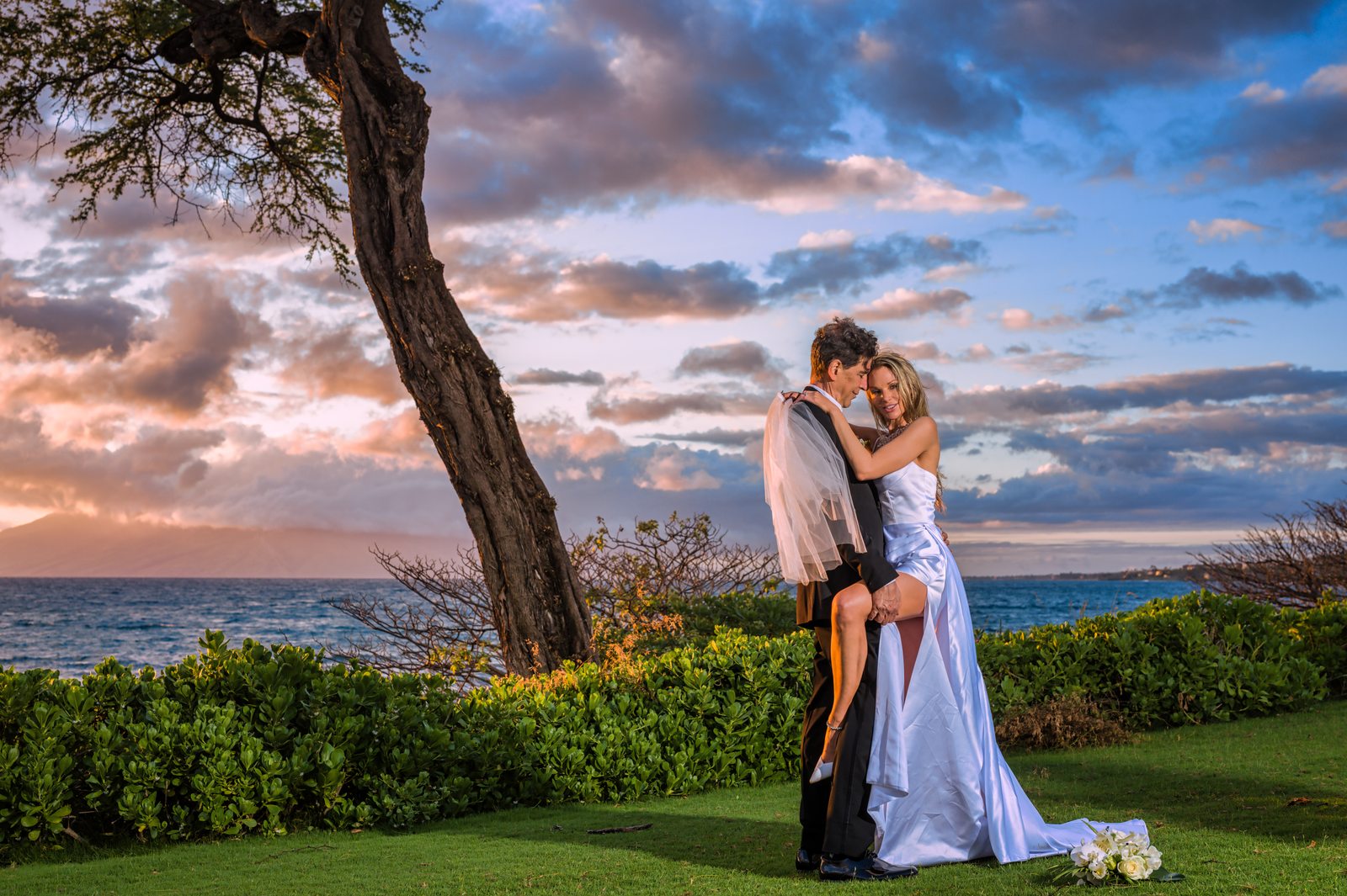 A couple exchanging vows beneath a floral arch at an oceanfront Maui wedding ceremony, backlit by the sun