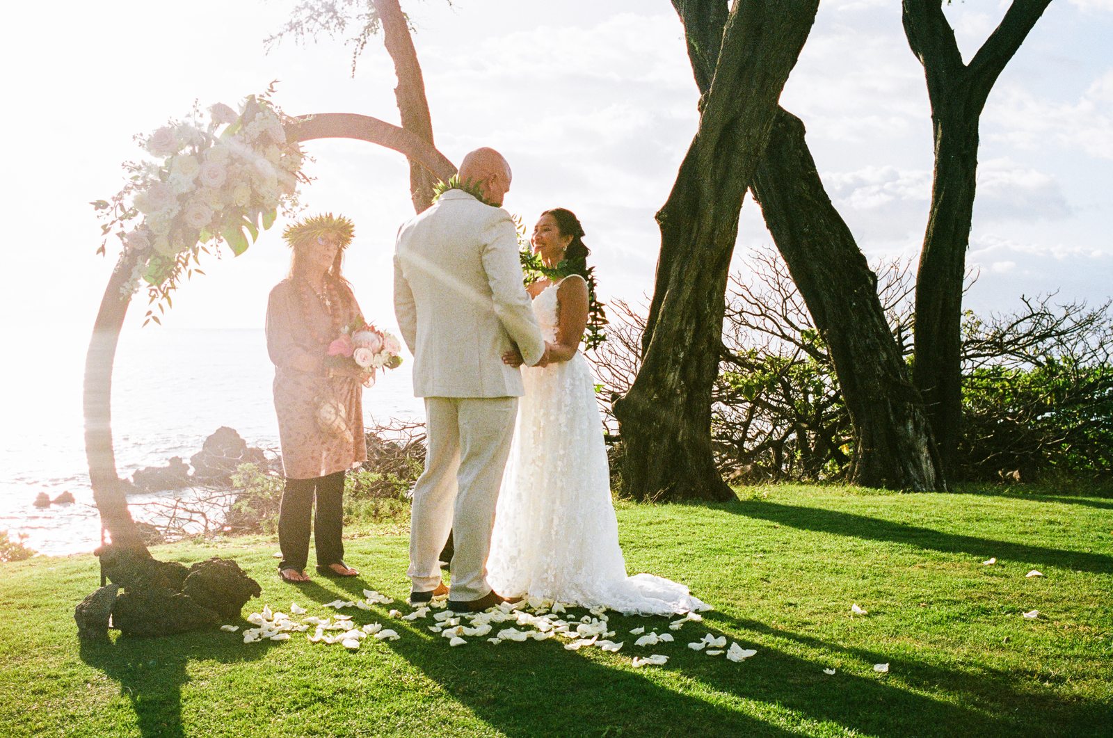 A bride and groom facing each other during their vows beneath a flower-covered arch in Maui