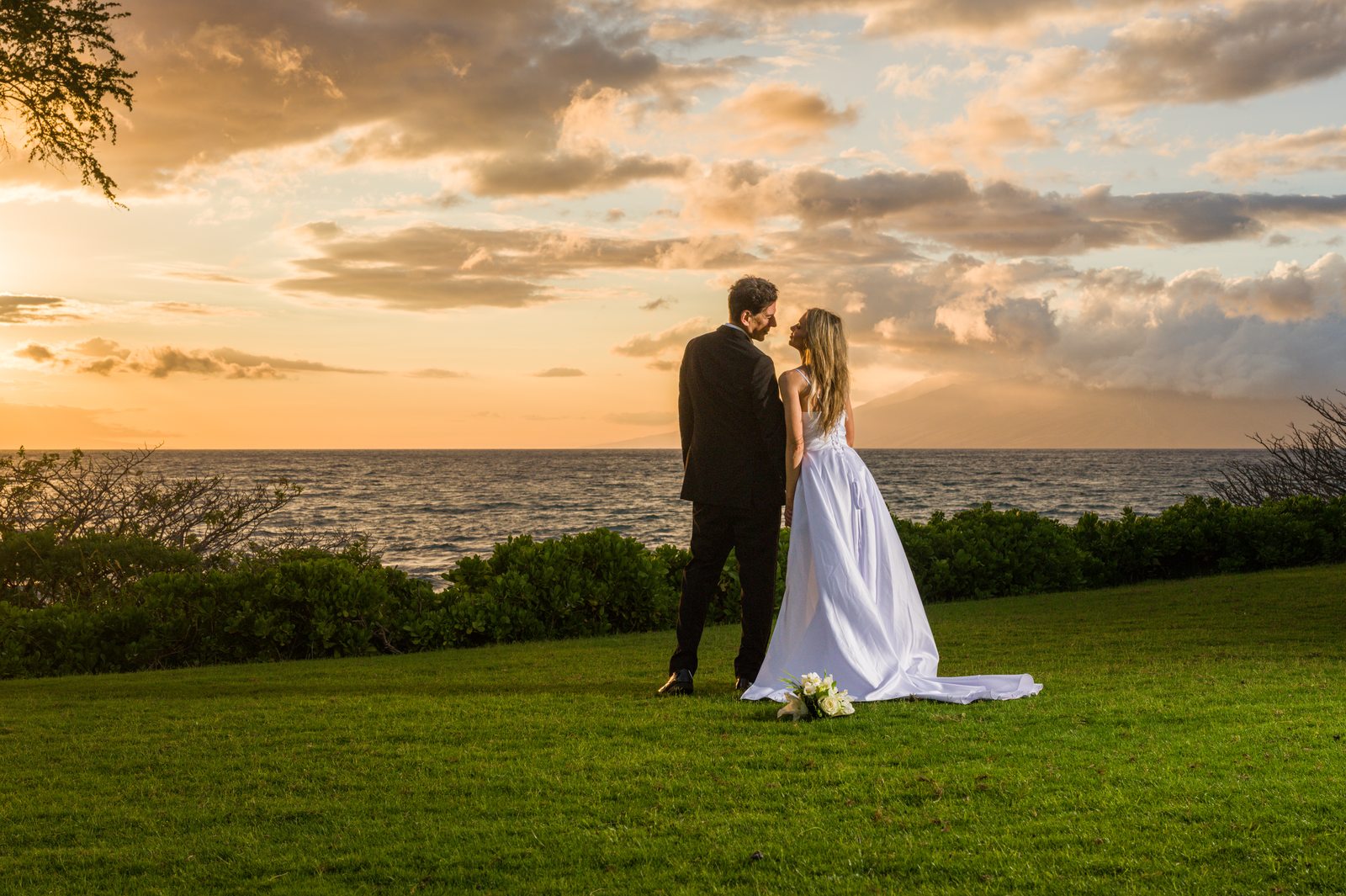 A bride and groom standing together on a Maui beach at golden hour, the bride holding a bouquet