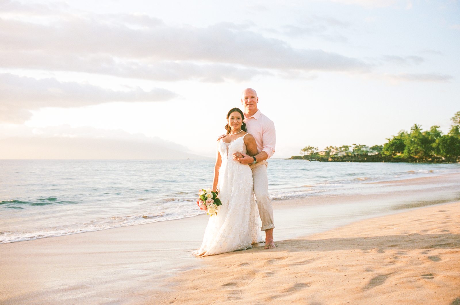 A bride and groom smiling together on a Maui beach at sunset, the bride holding her bouquet