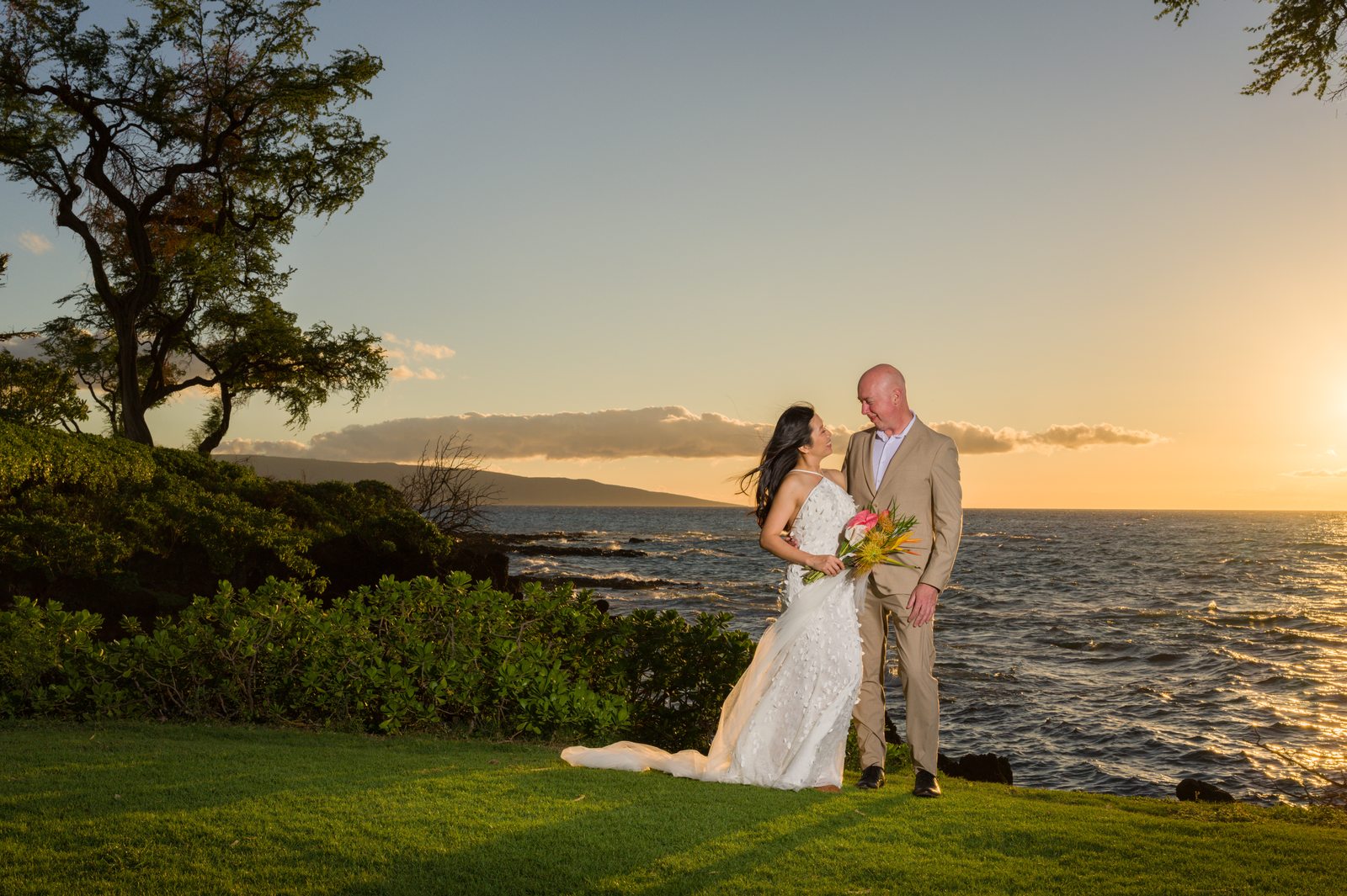 A bride and groom walking hand in hand across an oceanfront lawn in Maui at sunset
