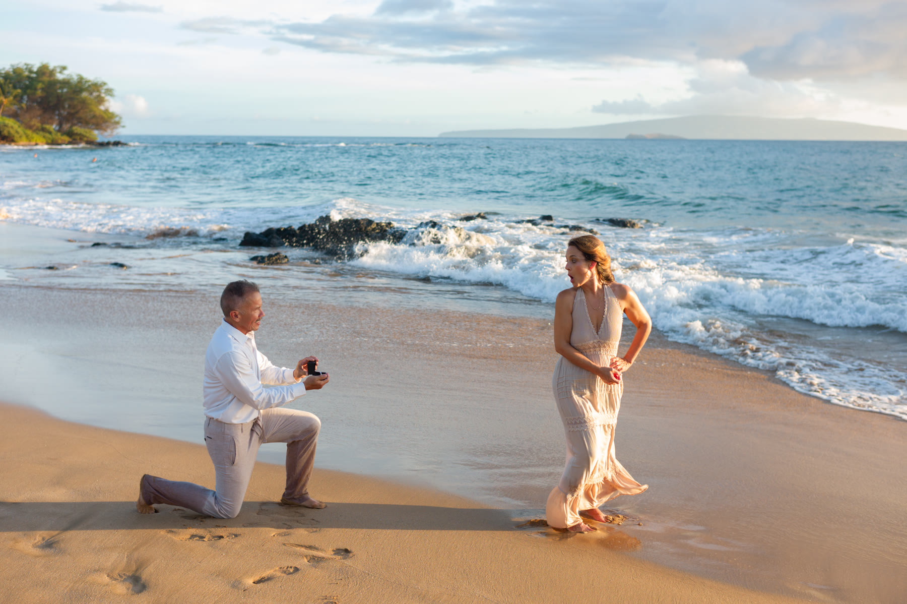 A woman turns and reacts as her partner kneels with an engagement ring on a Maui beach