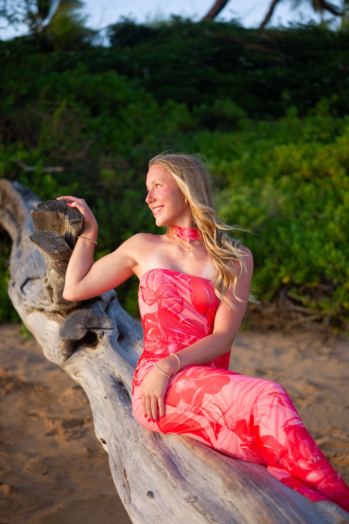 A senior seated on driftwood in a coral floral dress at golden hour in Maui