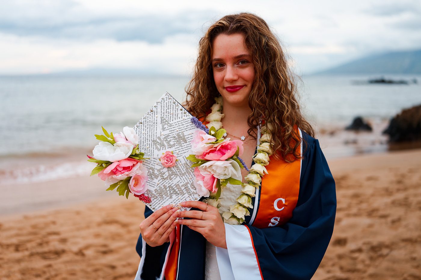 A graduate in cap and gown holding a decorated mortarboard on a Maui beach