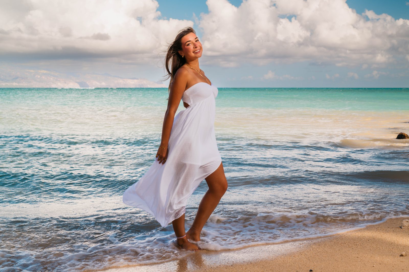 A senior in a white dress at the edge of turquoise water on a Maui beach