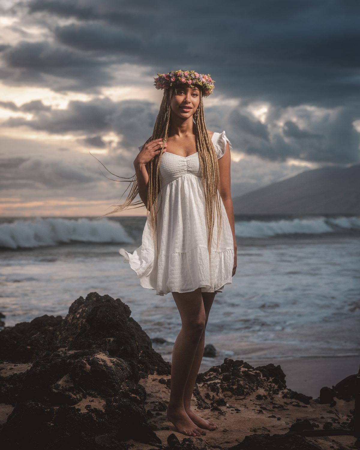 A senior in a white dress and flower crown beneath breaking waves at Mokapu Beach