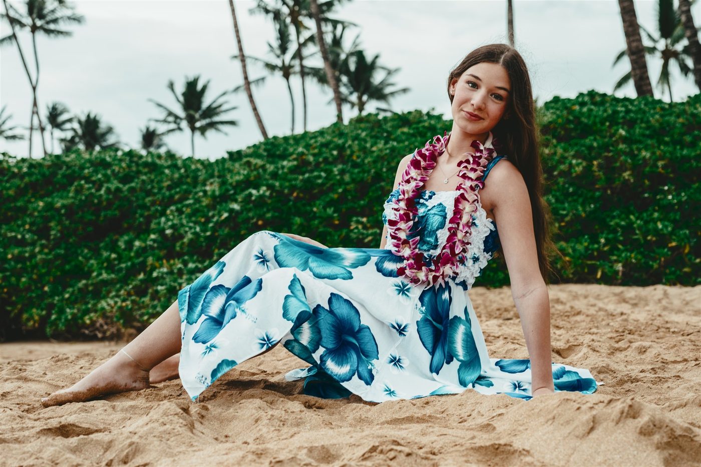 A senior seated on the sand in a blue hibiscus dress with an orchid lei at Mokapu Beach