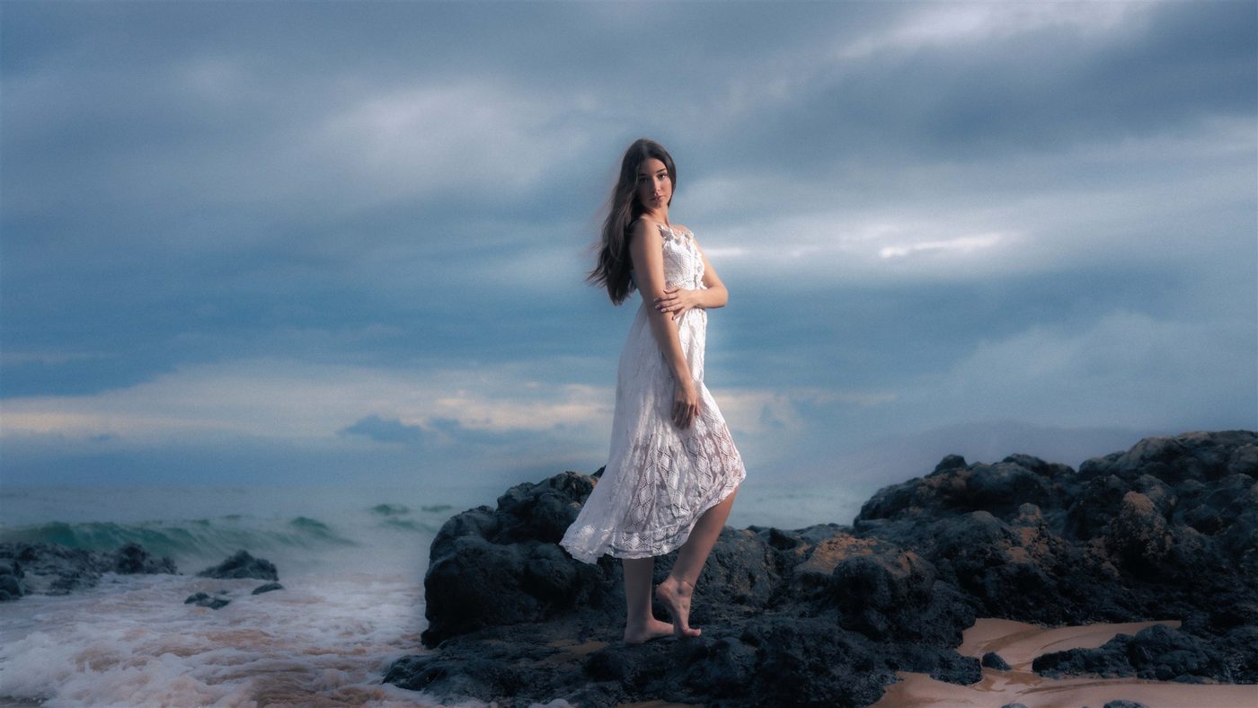 A senior in a white dress on the rocks beneath moody blue Maui clouds