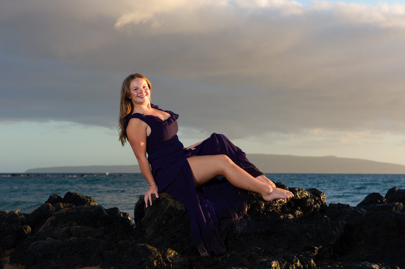A senior in a deep purple gown reclining on lava rock above the ocean in Maui