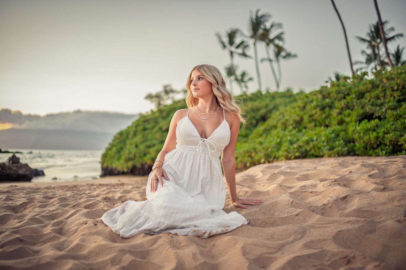 A senior seated on the sand at golden hour with palms behind her in Wailea