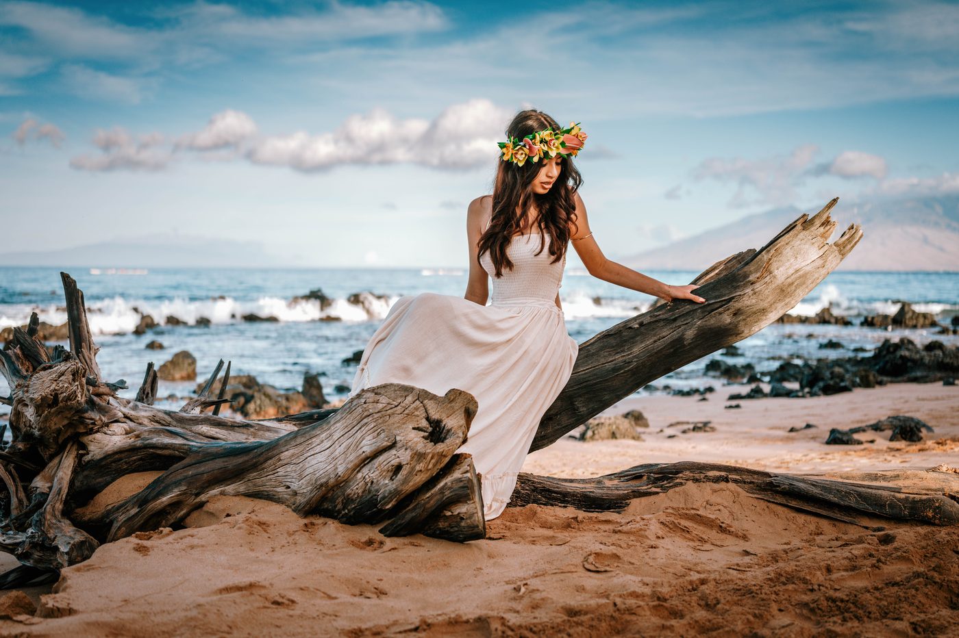 A senior in a flower crown seated on driftwood under a bright Maui morning sky