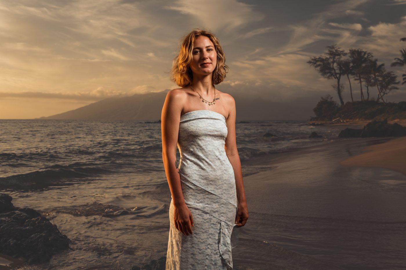 A senior in a white lace dress on the sand beneath a dramatic Maui sunset