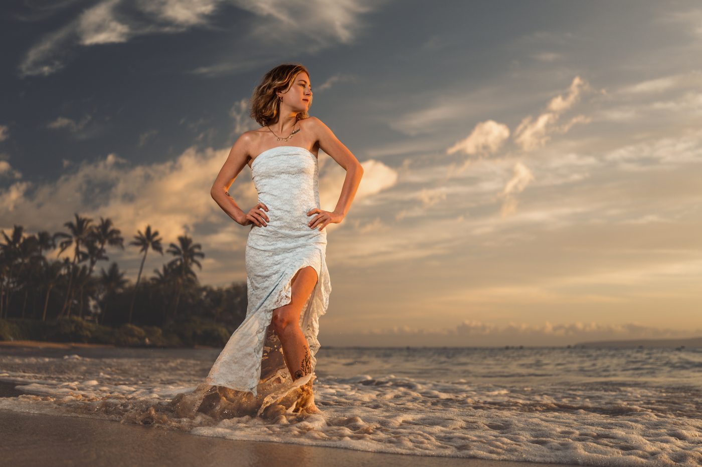 A senior standing in the shorebreak in a white lace dress in Maui