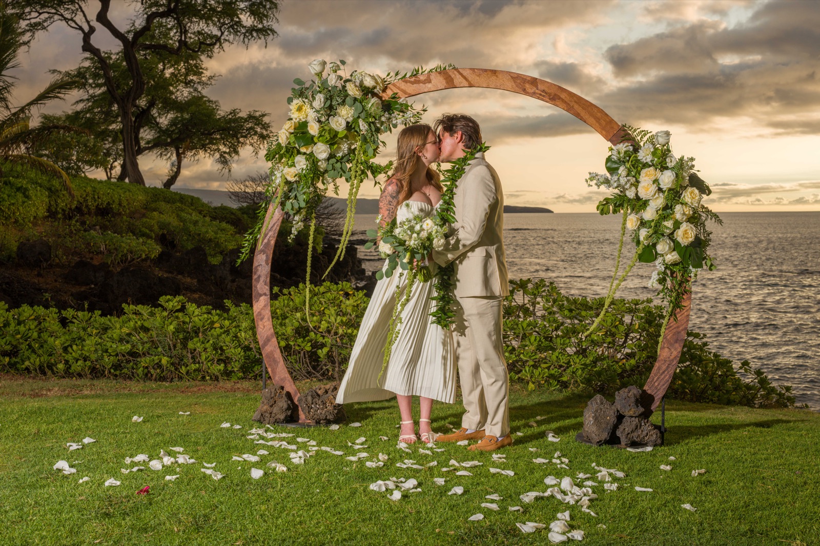 A newlywed couple kissing beneath a floral wedding arch overlooking the ocean in Wailea